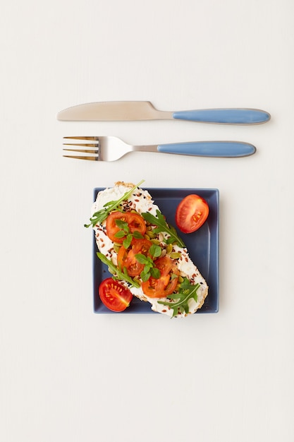 Zdjęcie widok z góry na minimalną kompozycję pojedynczej bruschetty z pomidorkami koktajlowymi i ziołami obok noża i widelca, zdrowe śniadanie i koncepcja diety