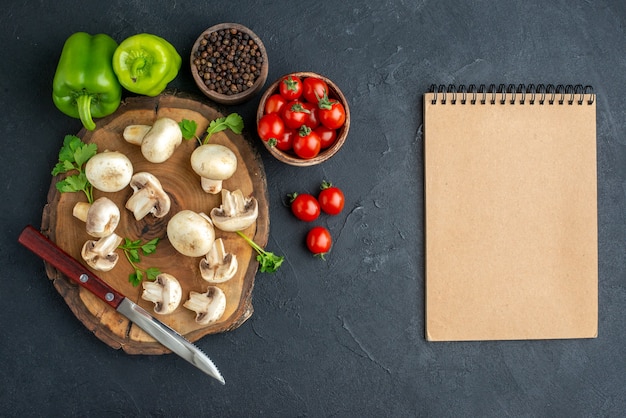 Widok z góry na nóż z surowych grzybów i zieleni na drewnianej desce biały ręcznik i notatnik spiralny ze świeżych warzyw na czarnym tle