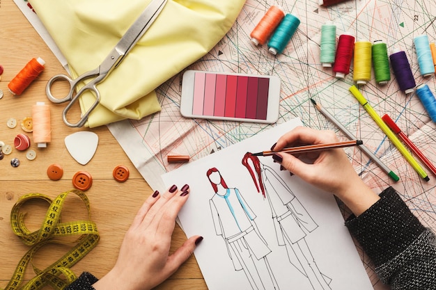 Zdjęcie widok z góry na projektanta mody w pracy. kobiece dłonie szkicują ubrania w jej kreatywnym miejscu pracy i używają smartfona z próbkami kolorów na ekranie, widok z góry