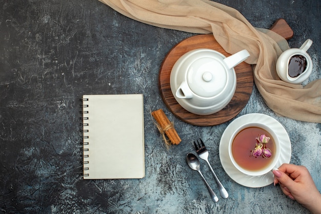 Widok z góry na rękę trzymającą filiżankę czarnej herbaty i czajnik na drewnianej tablicy notatnik cynamonowe limonki na lodowym tle