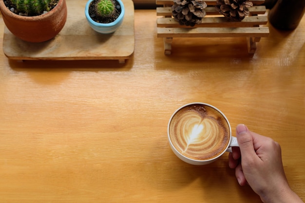 Widok z góry na rękę trzymającą filiżankę kawy latte art na brązowym drewnianym stole z kaktusową doniczką, miejsce na tekst for