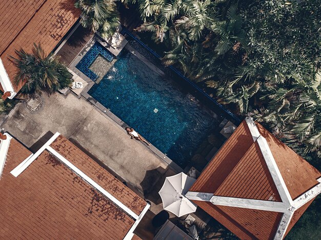Widok z góry na teren egzotycznego hotelu: dachy wyłożone terakotą, palmy, basen z błękitną wodą i młoda kobieta leżąca przy basenie; warkot.