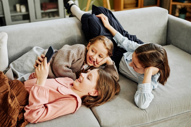 Widok z góry na trzy uśmiechnięte dziewczynki bawiące się razem na kanapie w domu i używające smartfona