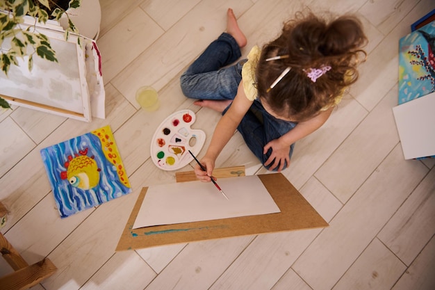 Widok z góry na uroczą dziewczynkę z Europy, cieszącą się malowaniem, siedzącą na podłodze przy drewnianej sztaludze na tle kolorowych, radosnych dzieci malujących obrazy Klasa plastyczna, kreatywność i edukacja