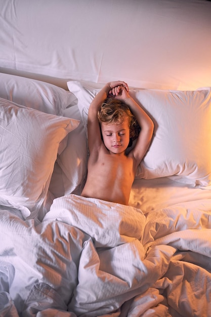 Widok z góry na uroczego chłopca bez koszuli z uniesionymi ramionami leżącego pod kołdrą w miękkim łóżku i śpiącego wieczorem w domu