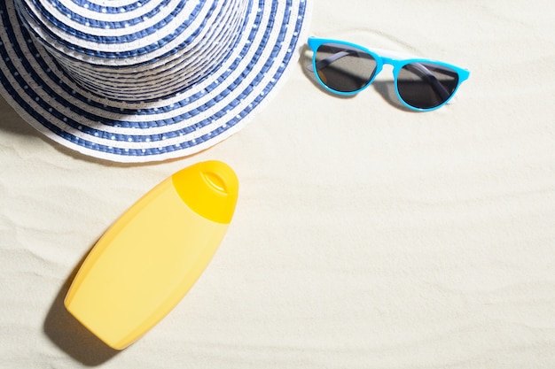 Widok z góry na żółtą kremową butelkę na piasku z kapeluszem i niebieskimi okularami przeciwsłonecznymi