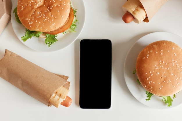 Widok z góry obraz śmieciowego jedzenia na stole smaczna kiełbasa w cieście i duże hamburgery inteligentny telefon z czarnym pustym ekranem miejsce na kopię reklamy lub tekstu promocyjnego