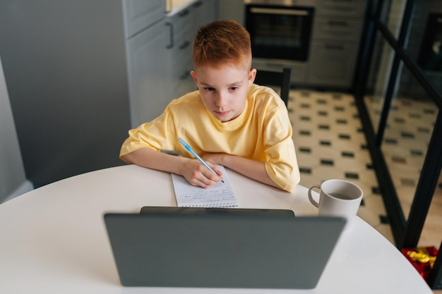 Widok z góry sprytnego rudowłosego ucznia robiącego notatki oglądającego lekcje online na laptopie z koncentracją w salonie