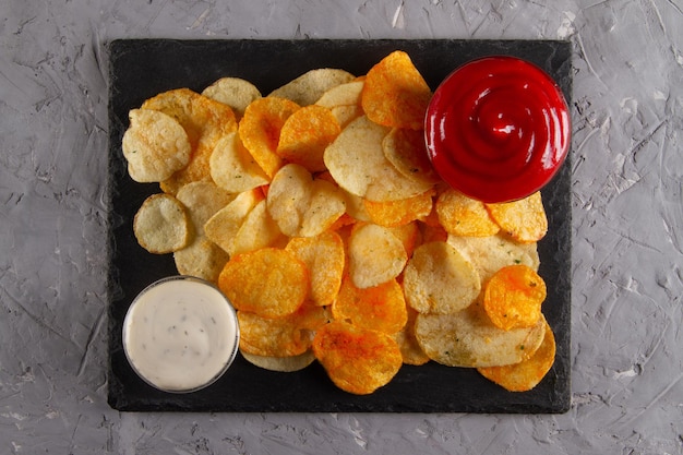 Widok z góry sterty chipsów ziemniaczanych z dwoma sosami do maczania na czarnej tabliczce do serwowania łupków na szarym plastrze
