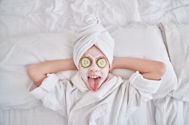 Zdjęcie widok z góry uroczego pozytywnego dzieciaka z ręcznikiem na głowie i maską na twarz z ogórkami relaksującymi podczas zabiegu pielęgnacji skóry i pokazującym język
