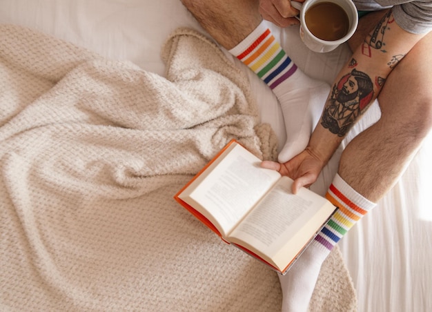 Widok z góry wytatuowanego mężczyzny w tęczowych skarpetkach pijącego kawę i czytającego książkę w łóżku rano