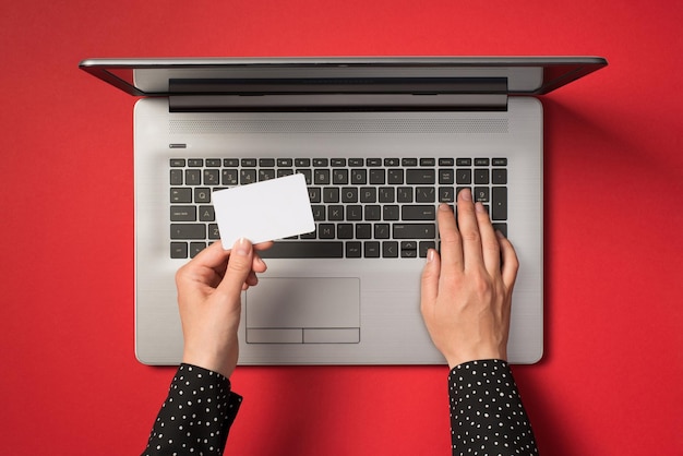 Widok z góry z pierwszej osoby przedstawiający dłonie piszące na klawiaturze laptopa i trzymające małą prostokątną białą kartkę na odizolowanym czerwonym tle z pustą przestrzenią
