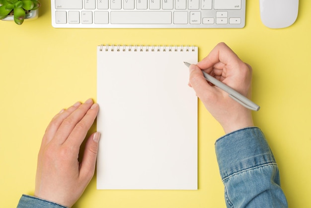 Widok z góry z pierwszej osoby przedstawiający kobiece dłonie trzymające długopis nad otwartym notatnikiem biała klawiatura myszy na izolowanym żółtym tle z copyspace