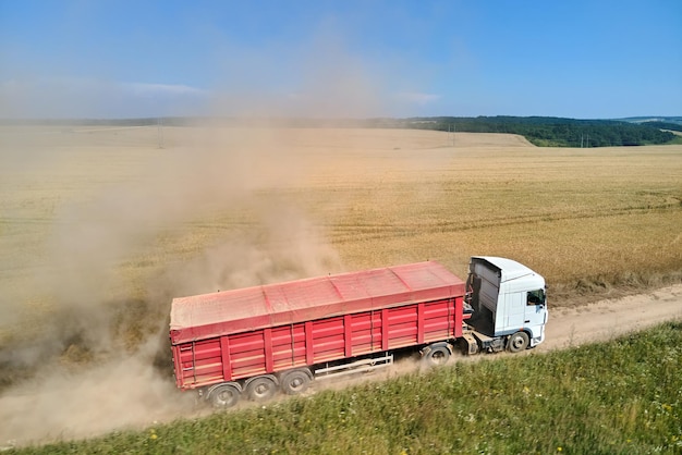 Widok z lotu ptaka ciężarówki jazdy na polnej drodze między polami pszenicy rolniczej, co powoduje dużo kurzu. Transport ziarna po zbiorze kombajnem w sezonie żniwnym