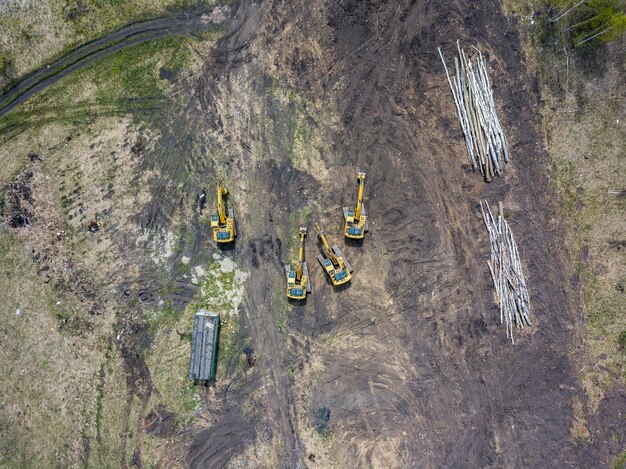 Widok z lotu ptaka czterech żółtych koparek gąsienicowych, które stoją na ziemi w pobliżu placu budowy i czekają na rozpoczęcie dnia roboczego, kopiąc ziemię w pobliżu powalonych drzew i drogi