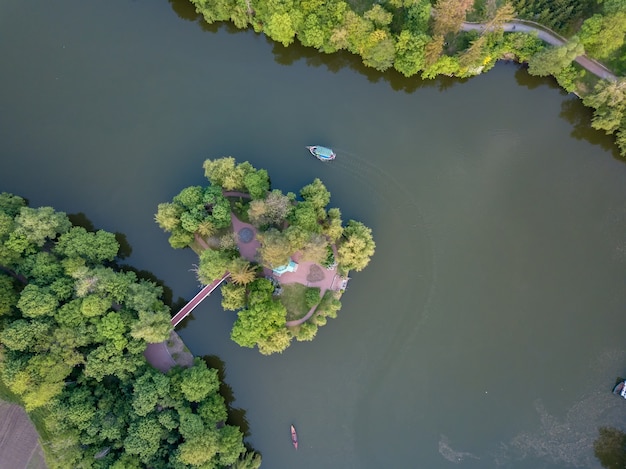 Widok z lotu ptaka na małe jezioro w środku lasu i wyspę z mostem wśród ogromnego zielonego lasu. Po wodzie płynie mała łódka. Zdjęcie drone