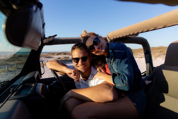 Widok z przodu białej pary w otwartym samochodzie, obejmującej i uśmiechającej się do kamery. Weekendowe wakacje na plaży, styl życia i wypoczynek.