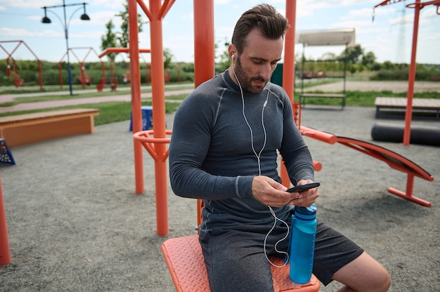 Widok Z Przodu Europejskiego Sportowca Noszącego Słuchawki, Trzymającego Telefon Komórkowy I Wskazującego Palcem Na Czarny Pusty Ekran Z Miejscem Na Kopię. Sportowiec Przesuwający Palcem Po Aplikacji Mobilnej Sprawdzający Bicie Serca