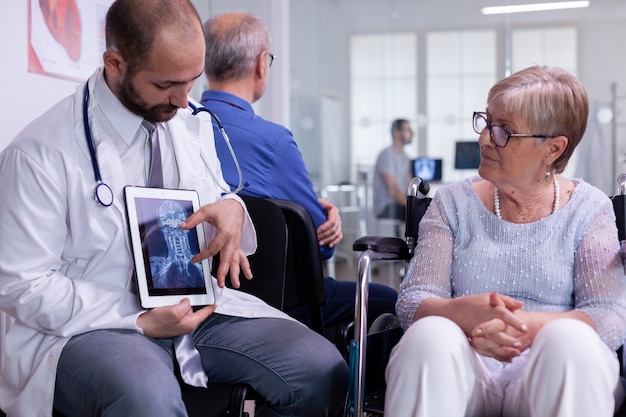 Widok z przodu lekarza rozmawiającego z niepełnosprawną starszą kobietą o leczeniu regeneracyjnym pokazującym radiografię za pomocą tabletu