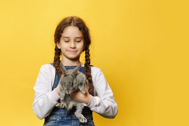 Widok z przodu małej słodkiej dziewczyny w dżinsach trzymającej prawdziwego szarego królika na białym tle żółtego