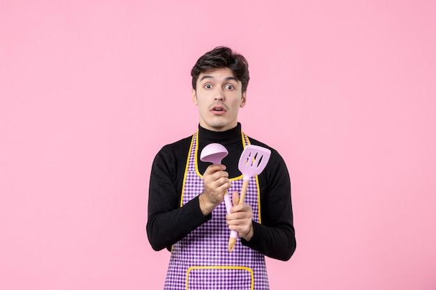 Widok z przodu młody mężczyzna w pelerynie trzymający łyżki na różowym tle zawód gotowanie posiłku praca pozioma ciasto kuchnia kolor jedzenie