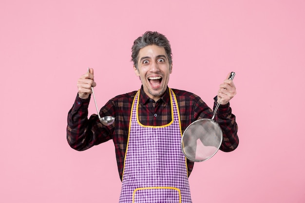 Zdjęcie widok z przodu młody mężczyzna w pelerynie z sitkiem i łyżką na różowym tle kuchnia kuchnia jedzenie pozioma zupa zawód kucharz jednolity pracownik mąż