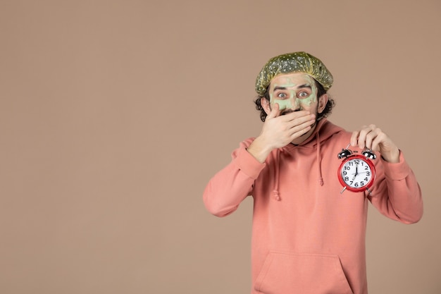 Zdjęcie widok z przodu młody mężczyzna z maską trzymający zegary na brązowym tle terapia uzdrowiskowa salon skóra pielęgnacja ciała twarzy