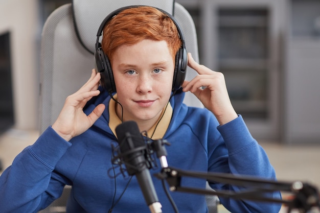 Widok z przodu portret rudowłosego nastoletniego chłopca mówiącego do mikrofonu i noszącego słuchawki podczas nagrywania podcastu lub przesyłania strumieniowego online
