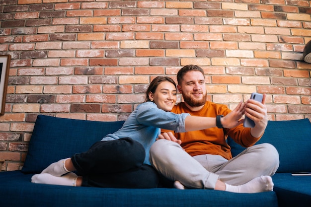 Widok z przodu szczęśliwej wesołej młodej pary, która ma czat online z kamerą internetową za pośrednictwem połączenia wideo na telefonie komórkowym, siedząc razem na wygodnej miękkiej sofie w przytulnym salonie. Pojęcie życia domowego.