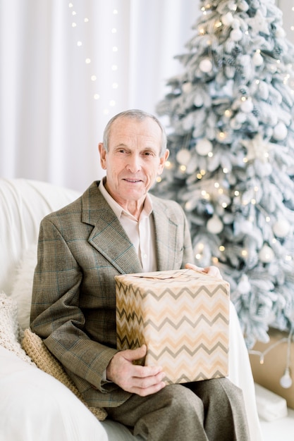 Widok z przodu szczęśliwy starszy mężczyzna w eleganckim garniturze siedzi w białym fotelu, trzymając obecne pudełko uśmiechając się do kamery w domu w domu na Boże Narodzenie