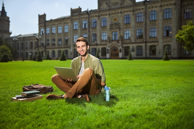 Widok z przodu wesołego studenta w swobodnym ubraniu, siedzącego ze skrzyżowanymi nogami na trawie