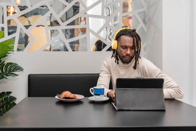 Widok z przodu współczesnego mężczyzny z dredami pracującego w kawiarni przy użyciu cyfrowego tabletu podczas słuchania muzyki przez żółte słuchawki