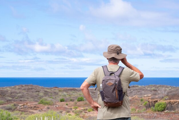 Widok z tyłu aktywnego starszego mężczyzny z plecakiem i kapeluszem podczas wędrówki na zewnątrz patrząc na horyzont Starszy mężczyzna na emeryturze i zdrowy tryb życia Błękitne morze i niebo w tle
