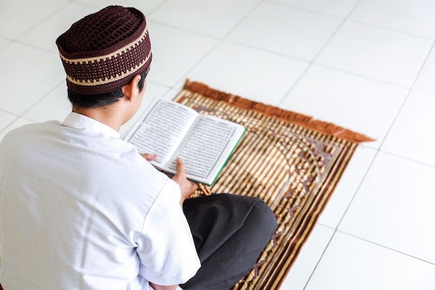 Widok z tyłu człowieka muzułmańskiego trzymającego i czytającego świętą księgę AlQuran na macie modlitewnej