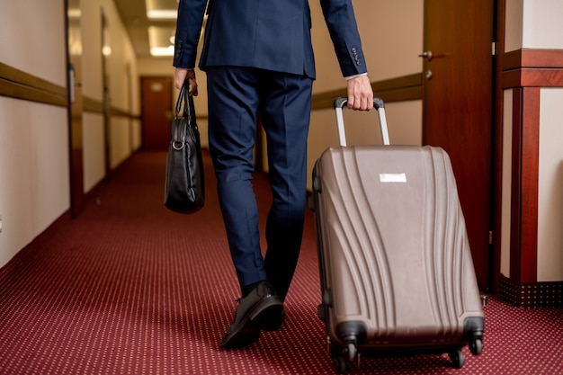 Widok z tyłu dolnej części eleganckiego biznesmena z walizką i torebką poruszającego się po korytarzu, wychodząc z hotelu
