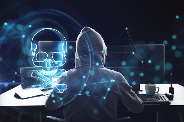Widok z tyłu hakera przy biurku za pomocą laptopa z cyfrowym hologramem niebieskiej czaszki na ciemnym tle Wirus Ransomeware i koncepcja zagrożenia pirackiego Podwójna ekspozycja