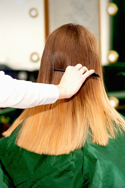 Widok z tyłu kobiecego fryzjera czesania długich włosów młodej blondynki klientki w salonie kosmetycznym
