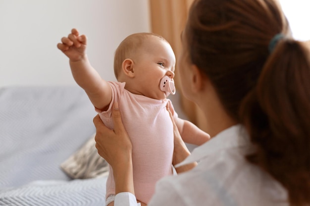 Widok z tyłu kobiety o ciemnych włosach Kobieta bawi się w domu ze swoją córeczką, trzymając dziecko z podniesionymi rękami, spędzając czas z uroczym dzieckiem w różowym body.