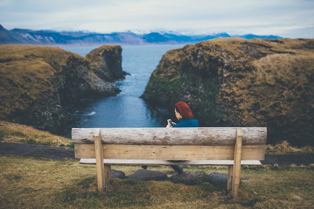 Widok z tyłu kobiety siedzącej na ławce i używającej smartfona w Islandii