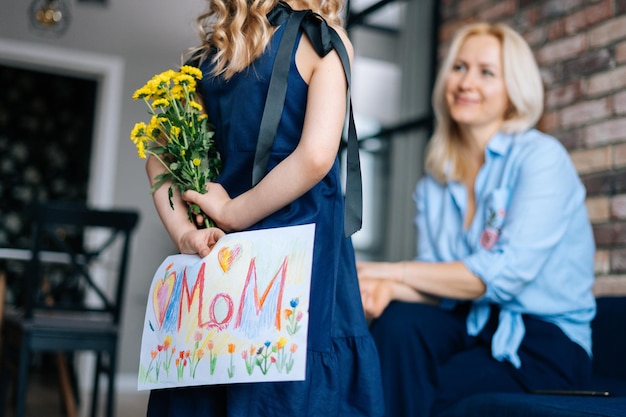 Widok z tyłu kręconej córki chowającej się za plecami niespodzianki dla matki. Dziewczyna daje mamie namalowany rysunek i bukiet kwiatów w domu w przytulnym salonie. Koncepcja szczęśliwy dzień matki.