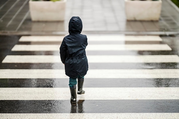 Widok z tyłu małego chłopca przechodzącego przez przejście dla pieszych na drodze w deszczową pogodę