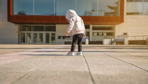 Widok z tyłu małej dziewczynki z kapturem patrząc na jej nowe trampki na placu miejskim w słoneczny dzień