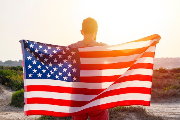 Widok z tyłu mężczyzny trzymającego macha flagą amerykańską USA przed zachodem słońca na zewnątrz nieba. Dzień Niepodległości Stanów Zjednoczonych Ameryki. Koncepcja amerykańskich patriotycznych ludzi
