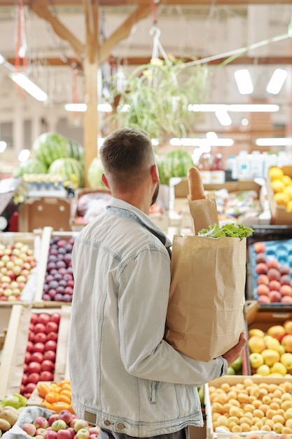 Widok z tyłu młodego mężczyzny w lekkiej dżinsowej kurtce trzymającego papierową torbę i wybierającego soczyste owoce na targu rolniczym