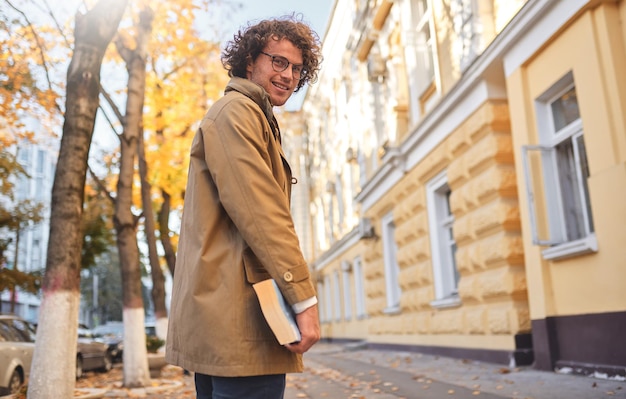 Widok z tyłu młodego mężczyzny w okularach pozuje z książką na zewnątrz Student collegu niosący książki w kampusie na jesiennej ulicy Uśmiechnięty facet nosi okulary i kręcone włosy, czyta książki na zewnątrz