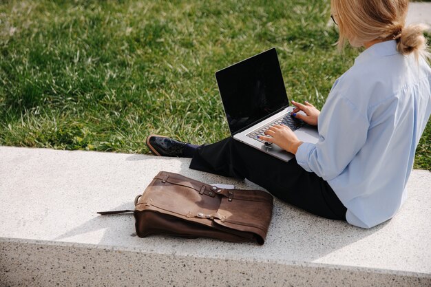 Widok z tyłu młodej blondynki w swobodnym stroju za pomocą nowoczesnego laptopa do pracy zdalnej, siedząc w ciepły dzień na świeżym powietrzu. Pojęcie ludzi i technologii.