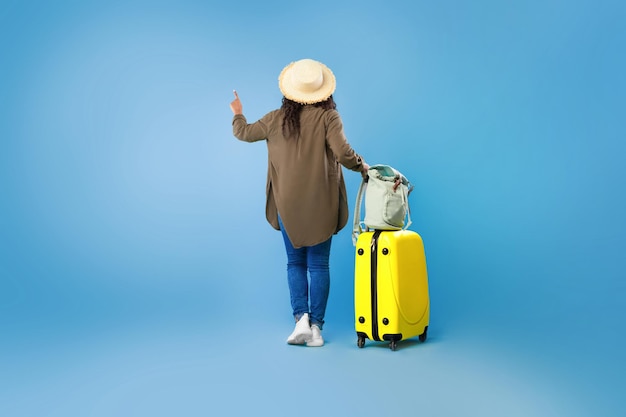 Widok z tyłu młodej czarnej kobiety z bagażem wskazującym wybór wycieczki turystycznej na niebieskim tle studia