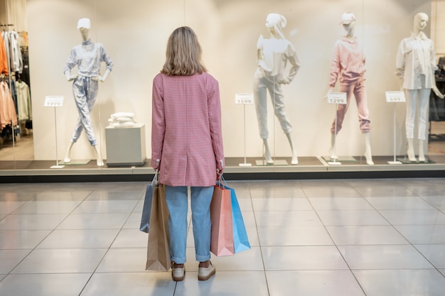 Zdjęcie widok z tyłu młodej kobiety trzymającej torby na zakupy i patrzącej na witrynę sklepu w centrum handlowym