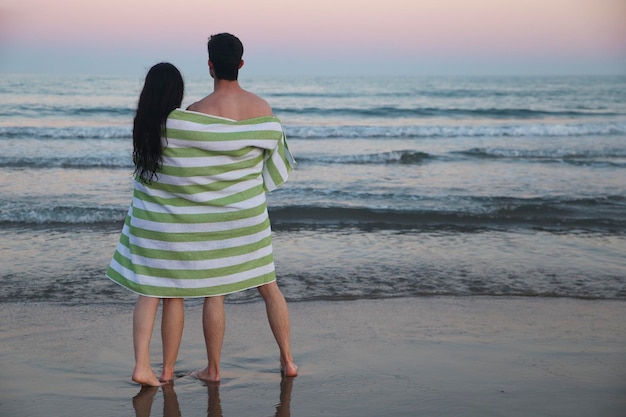 Widok z tyłu młodej pary na plaży owiniętej razem w ręcznik oglądając zachód słońca