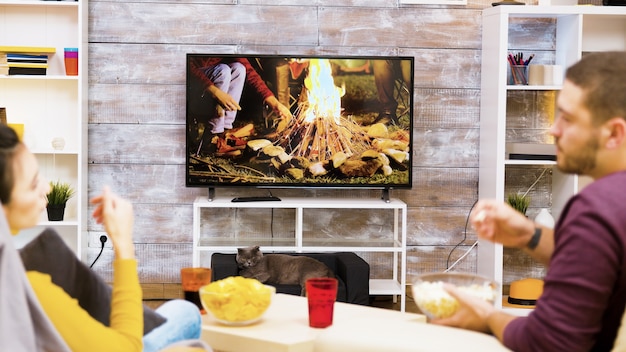 Widok z tyłu młodej pary relaks na krześle przed telewizorem podczas jedzenia frytek i popcornu. Kot przed telewizorem.